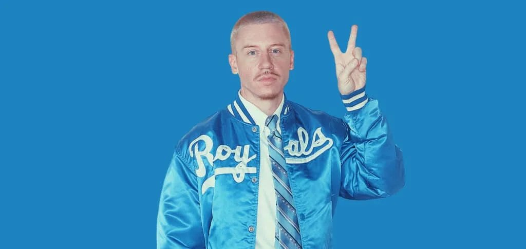 A person wearing a shiny blue jacket with the word 'Royals' printed in white, holding up a peace sign against a solid blue background.