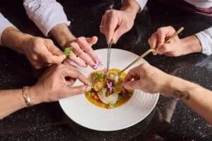 Several hands with utensils carefully plating an intricate dish garnished with flowers and herbs on a white plate.