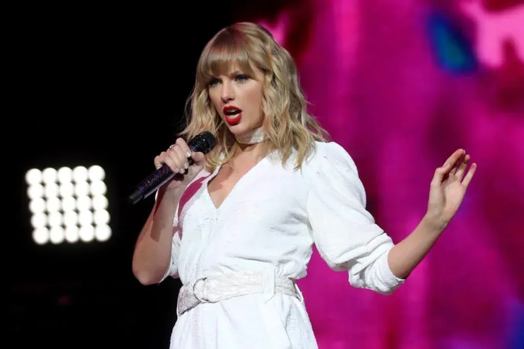 A woman with wavy blonde hair and bold red lipstick sings into a microphone on stage, wearing a white outfit with a matching belt.