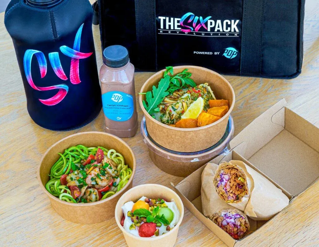 A variety of healthy meals in cardboard containers, including zoodles with shrimp, a salad with lemon, sweet potato, and a burrito cut in half. There is a chocolate drink bottle and a black lunch bag labeled 'The Six Pack Revolution.' A black water bottle with pink and blue graffiti-style lettering sits nearby on a wooden surface.