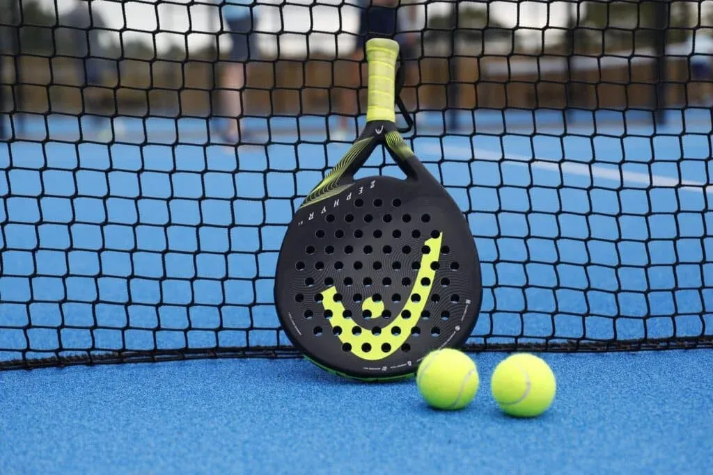 A black padel racket with a yellow design resting against a net on a blue court, accompanied by two yellow tennis balls.
