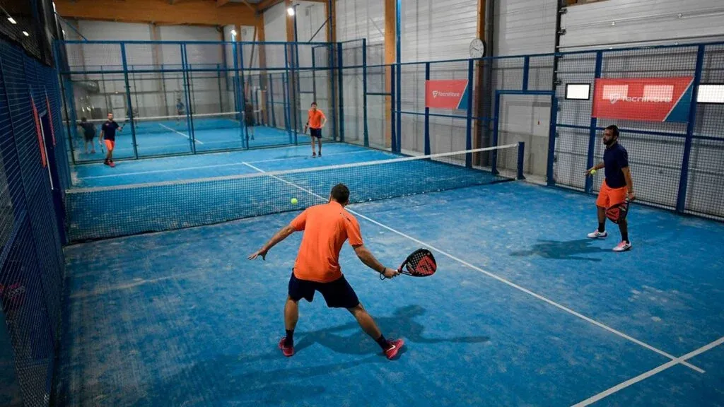 Four people playing paddle tennis on an indoor blue court, with two players on each side of the net.
