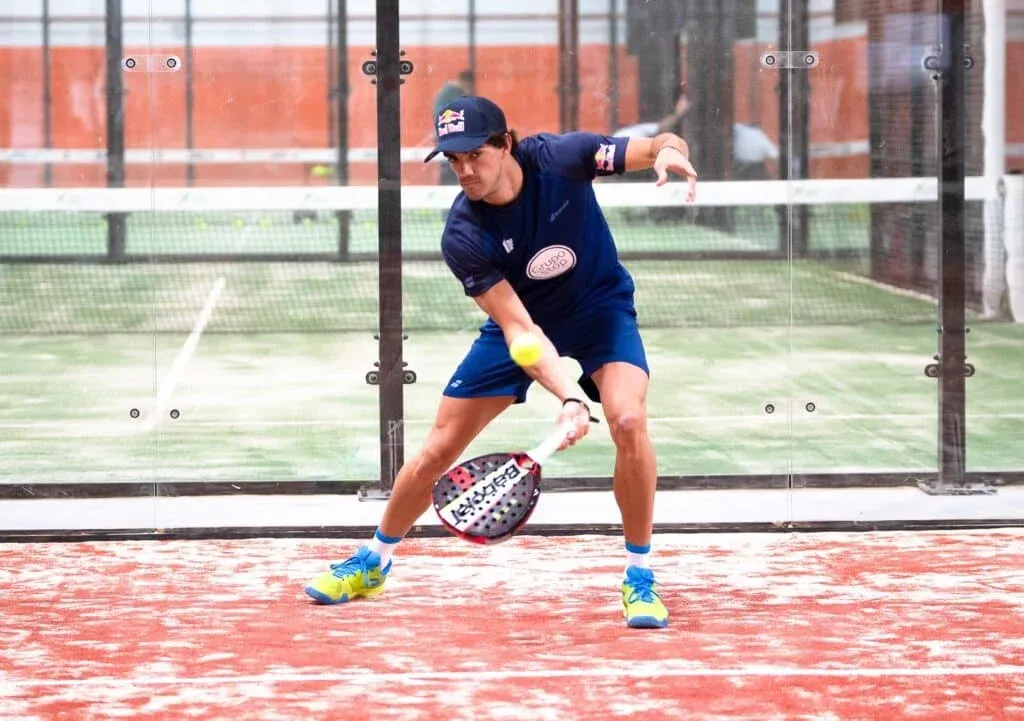 A person wearing a blue outfit and a cap playing paddle tennis, preparing to hit the ball with a paddle on a court surrounded by glass walls.