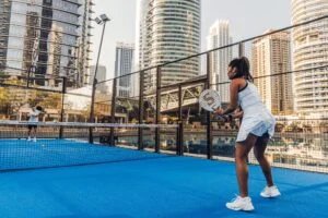Two people playing padel on a blue court surrounded by a high fence, set against a backdrop of tall modern buildings.