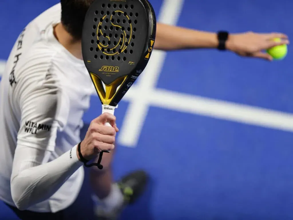 A person playing padel tennis, holding a black and yellow padel racket in the foreground, with the court's blue surface and white lines visible in the background.