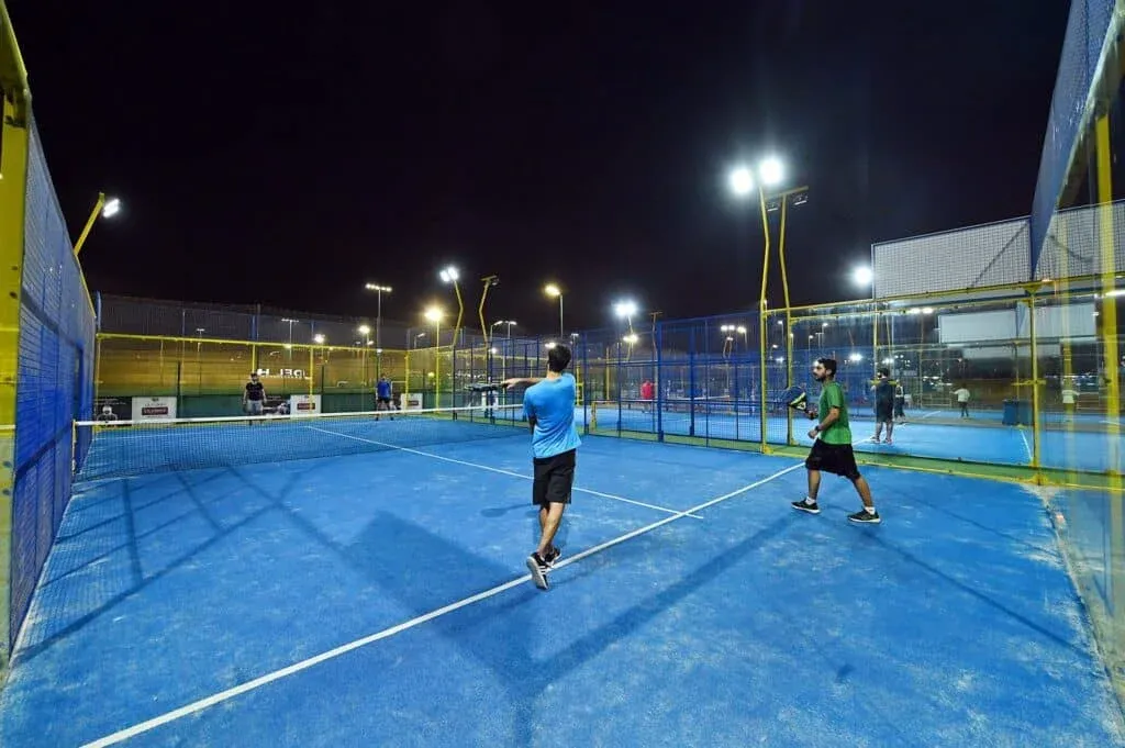 Two men playing padel on an illuminated blue court at night, surrounded by high fences, with other courts and players visible in the background.