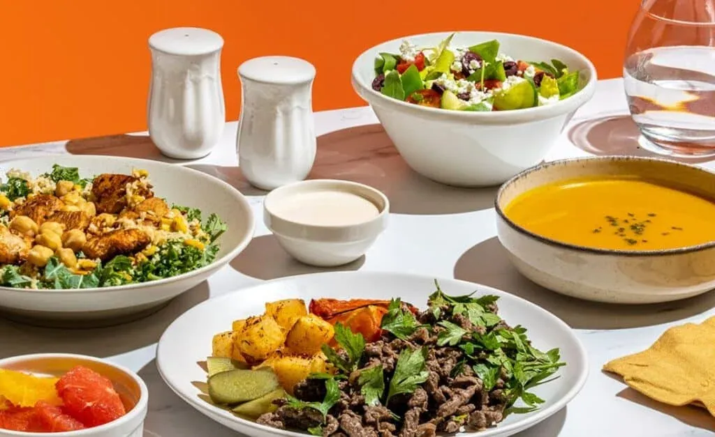 A variety of dishes on a table including a salad with sliced cucumbers, tomatoes, and feta cheese, a bowl of chickpea salad with kale, a plate of beef stir fry with roasted potatoes, a bowl of creamy orange soup garnished with herbs, a small dish of sauce, and a cup of fruit segments. The table is set against an orange background with a glass of water and salt and pepper shakers present.