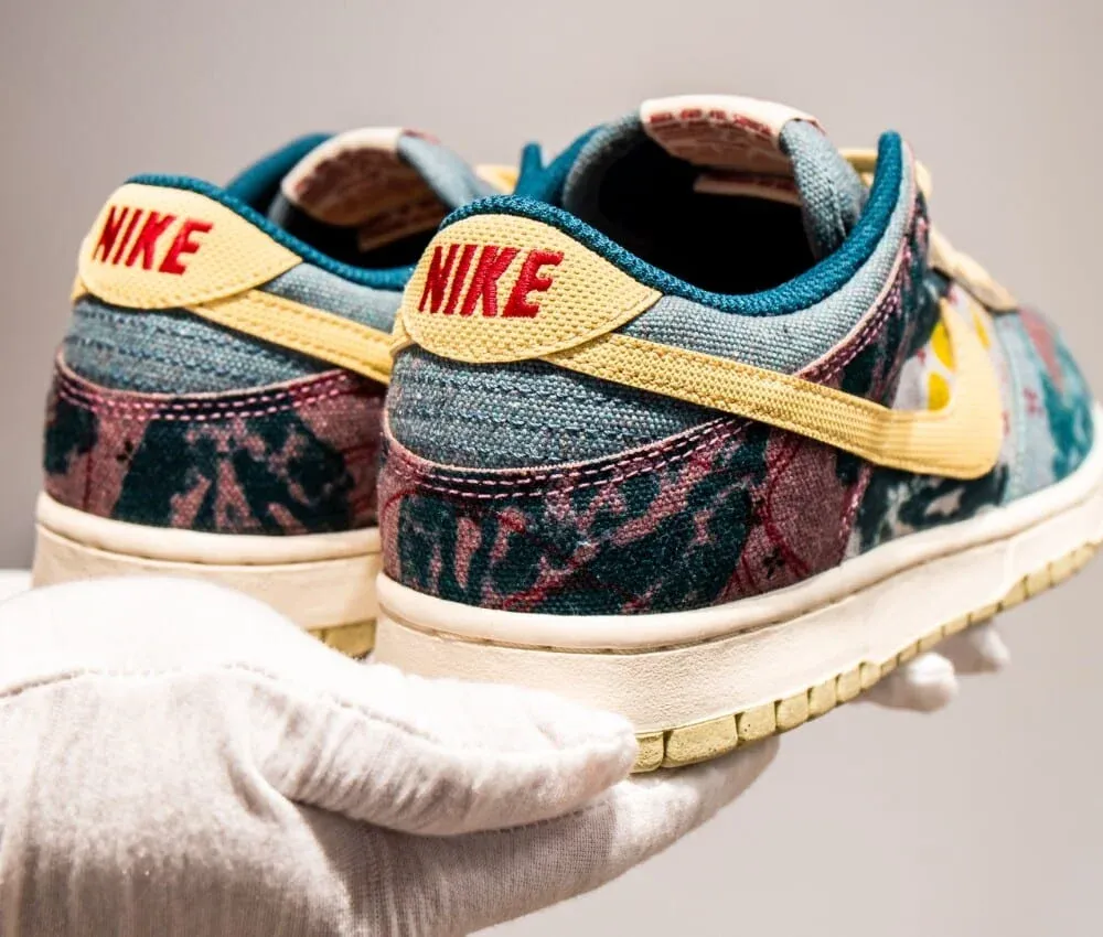 A pair of colorful Nike sneakers with a patchwork pattern in teal, maroon, and cream, held by a gloved hand. The sneakers feature a yellow Nike logo and white soles.