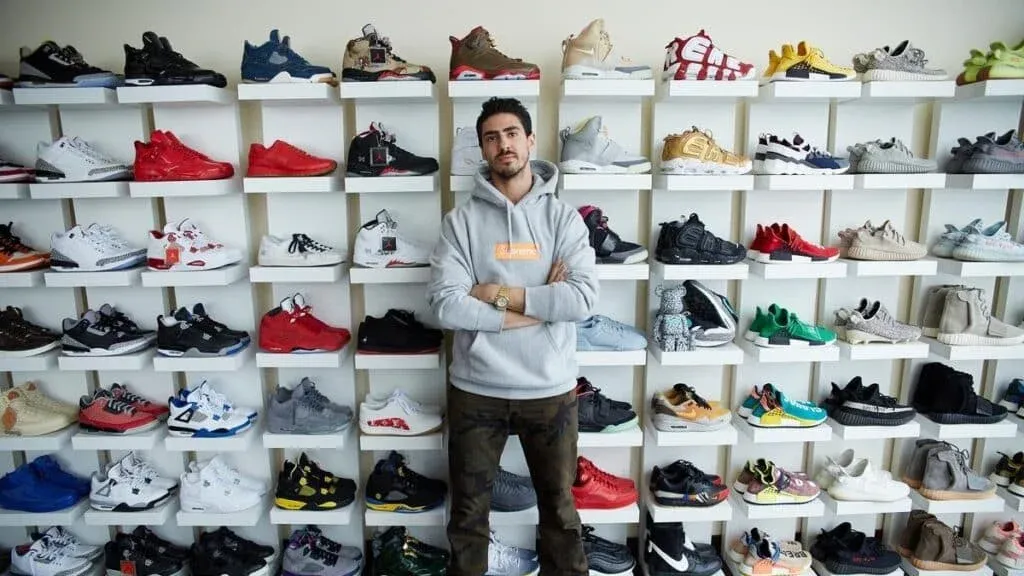 A person in UAE Sneaker Culture standing in front of shelves filled with a large collection of diverse sneakers, wearing a gray hoodie and crossing their arms.
