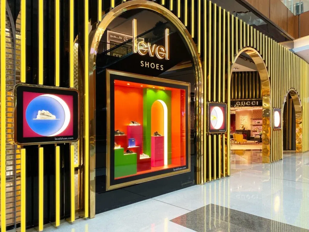A opulent shoe store display with the name 'Level Shoes' prominently displayed above a brightly lit showcase window. The window exhibits footwear against a lively, multicolored backdrop. The surrounding structure features gleaming gold accents and striped patterns. Adjacent is a peek into another luxurious retail space.