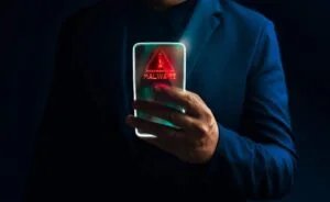 A person in a dark suit holds a smartphone displaying a glowing red malware warning with an exclamation mark inside a triangle.