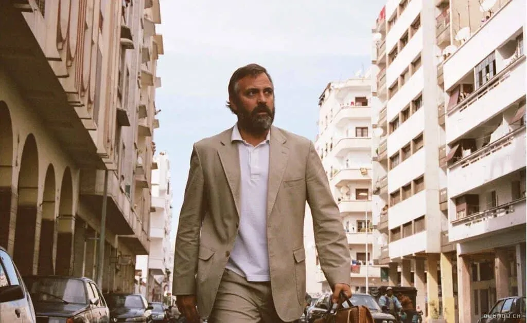 A man with a beard walks down a street between tall buildings, wearing a beige suit and carrying a brown leather bag.