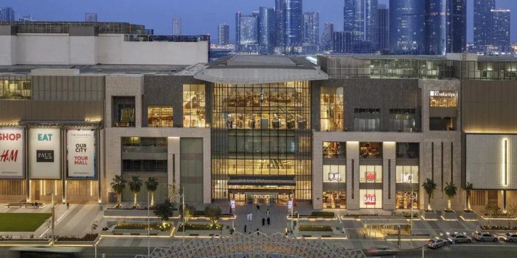 A large, modern shopping mall featuring illuminated signs for various stores such as H&M and Kinokuniya, with a city skyline in the background. The mall's facade has large glass windows and is surrounded by neatly landscaped grounds with trees and a road in front featuring parked cars.