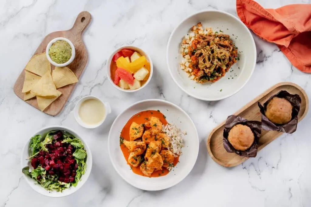 An assortment of dishes on a marble surface, including a bowl of leafy salad with beets, a small board with tortilla chips and guacamole, a cup of fruit salad, a sauce dish, a plate with orange-sauced chicken over rice, a dish of meat on vegetables, and two muffins on a wooden tray.