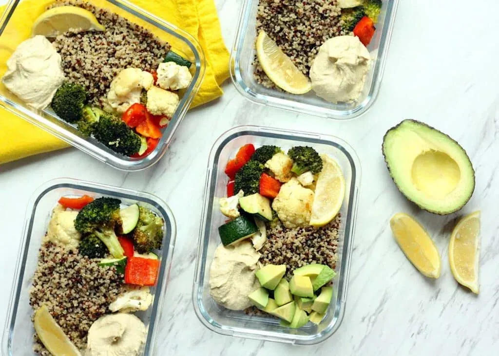 Four meal prep containers filled with quinoa, roasted broccoli, cauliflower, zucchini, red bell peppers, hummus, and lemon wedges, with an avocado and more lemon slices on the side.