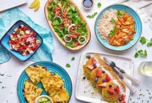 An array of vibrant dishes on a table, including a plate of pancakes with berries, a colorful salad with greens and onions, a bowl of rice with vegetable curry, a blue bowl with chopped fruits, and a plate of quesadillas with corn and beans, accompanied by a small dish of guacamole.