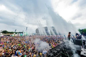 A large crowd at a festival is enjoying a water spray show on a stage, with many people raising their hands and wearing colorful attire under a cloudy sky.