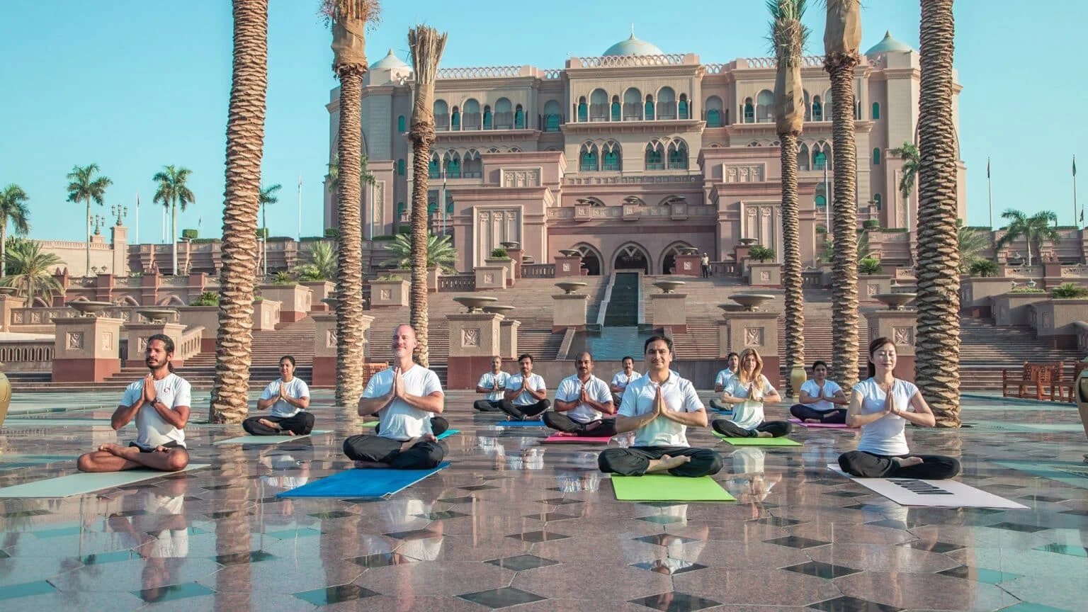 A group of people practicing yoga on colorful mats in front of an ornate building with palm trees, under a clear blue sky.