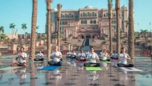 A group of people practicing yoga on colorful mats in front of an ornate building with palm trees, under a clear blue sky.