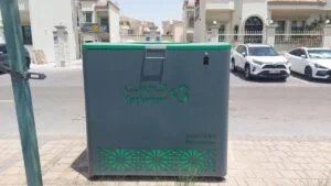 A large gray recycling bin with a green top, labeled "Tadweer Recyclables" in both Arabic and English, situated on a street with parked cars and buildings in the background.