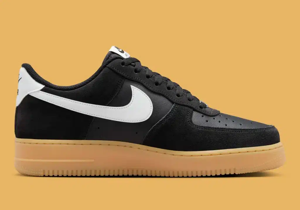 A black suede sneaker with a white swoosh logo and a gum-colored sole, set against a mustard yellow background.