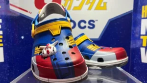 A pair of Crocs styled in a red, blue, and yellow design with a small figurine decoration attached to one shoe, displayed against a branded backdrop.