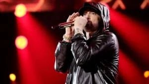 A man in a black leather jacket and hat performs on stage, holding a microphone with a background lit in red lights.