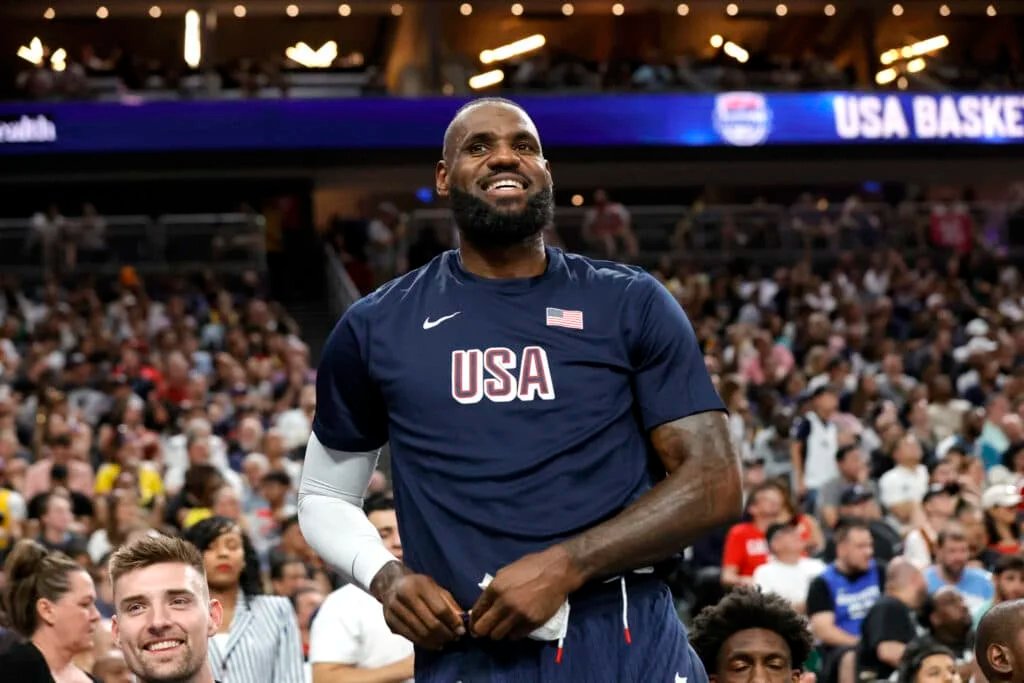 A basketball player wearing a navy blue USA shirt smiling in front of a crowded arena.