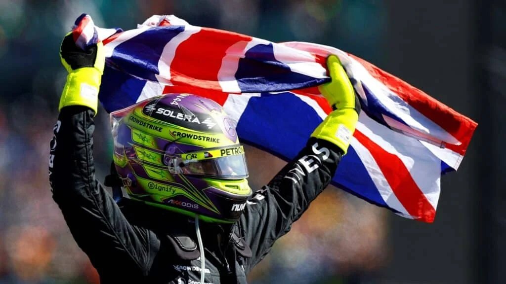 A person in racing gear with a colorful helmet and neon gloves holding a British flag overhead against a blurred background.