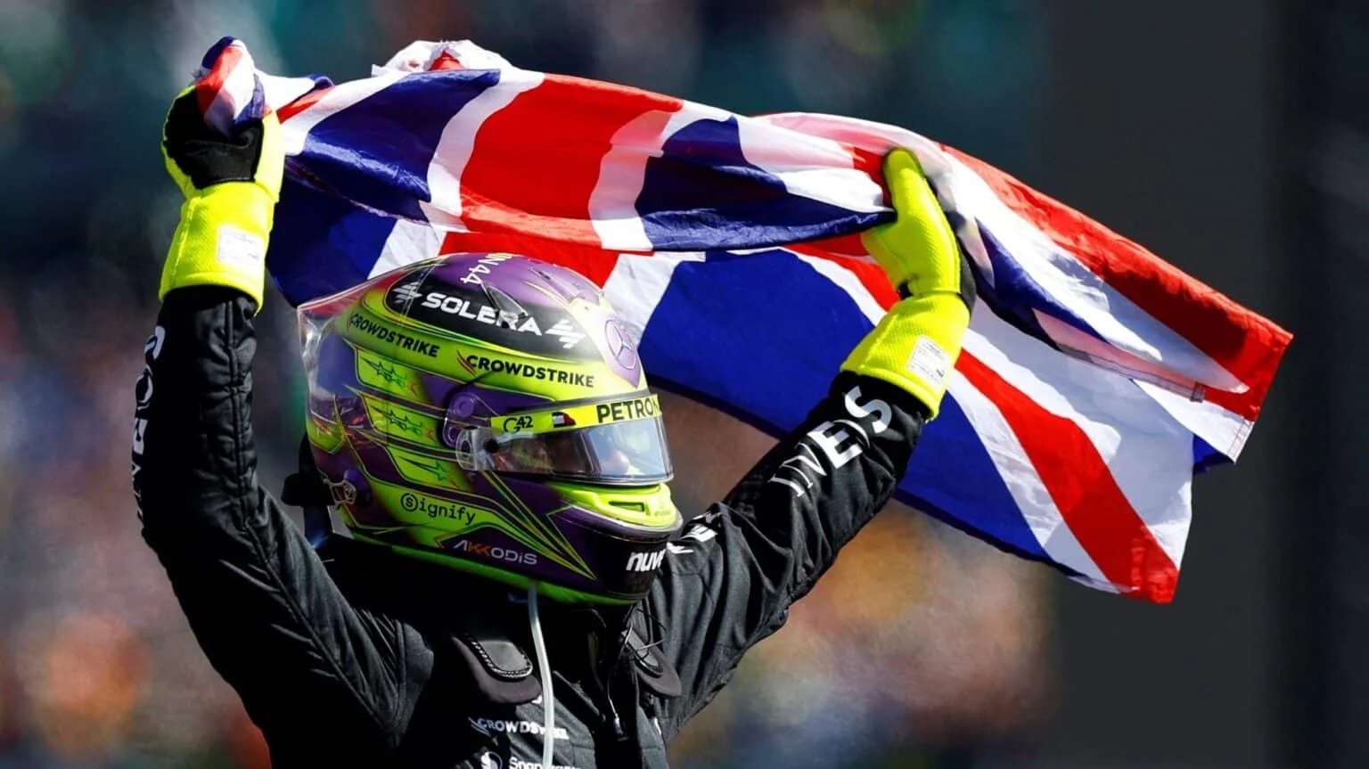 A person in racing gear with a colorful helmet and neon gloves holding a British flag overhead against a blurred background.