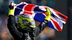 A person in racing gear with a colorful helmet and neon gloves holding a British flag overhead against a blurred background.