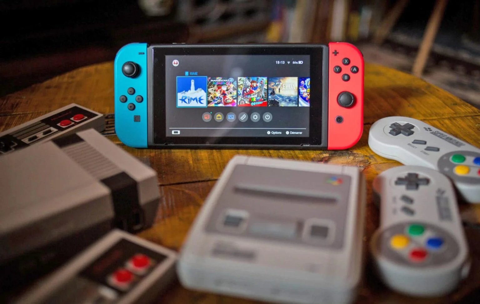 A Nintendo Switch with blue and red Joy-Con controllers, displaying a game menu on the screen, is placed on a wooden table surrounded by retro gaming consoles and controllers, including a NES and SNES.
