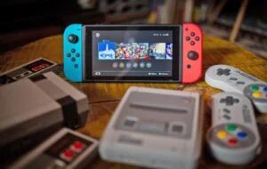 A Nintendo Switch with blue and red Joy-Con controllers, displaying a game menu on the screen, is placed on a wooden table surrounded by retro gaming consoles and controllers, including a NES and SNES.