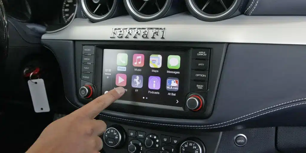 A hand touches the display of a car's infotainment system featuring various app icons, with the brand name 'Ferrari' above it.