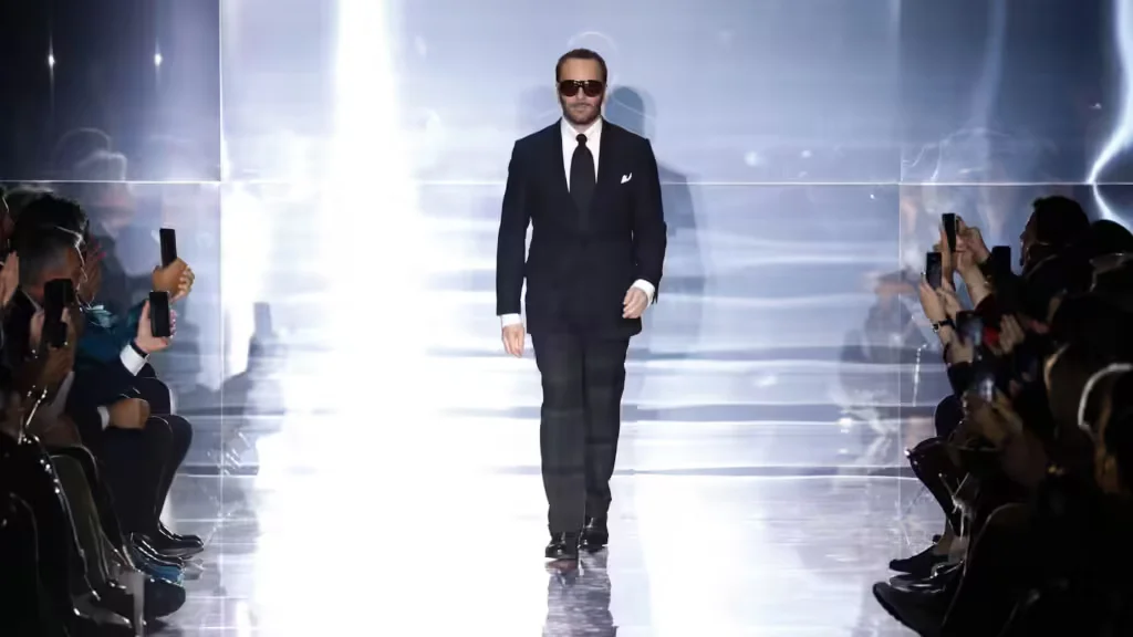 A man in a black suit and sunglasses walks confidently down a runway with bright lighting, surrounded by a crowd holding up smartphones to take pictures.