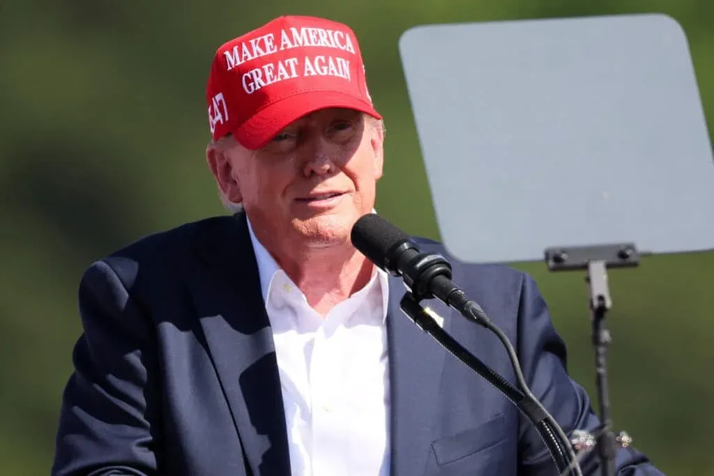 A man wearing a red hat with the slogan 'Make America Great Again' is speaking into a microphone, standing in front of a teleprompter.