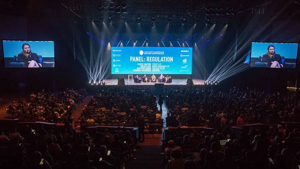 A large audience seated in a dimly lit auditorium watches a panel discussion on stage. The stage features a large screen displaying the topic