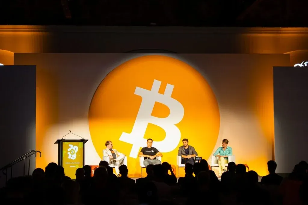 A panel discussion at a Bitcoin conference featuring four people seated on stage in front of a large Bitcoin logo. The stage is illuminated in orange and white, with an audience visible in the foreground.