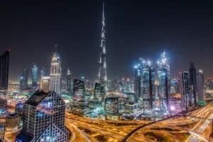 Nighttime view of a modern cityscape featuring brightly lit skyscrapers, with a prominent tall tower in the center, surrounded by illuminated roads and buildings.