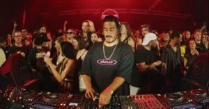 A DJ performing at a crowded event, surrounded by people under red lighting. The DJ is wearing a black shirt with the word 'deus' on it and has headphones around his neck. A DJ console with controls and turntables is visible in the foreground.