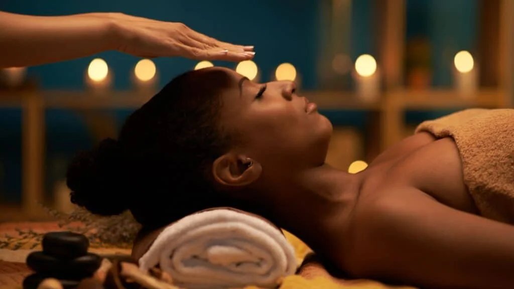 A woman lying down with her head resting on a rolled towel, eyes closed in relaxation. A hand gently hovers above her forehead, suggesting a massage or healing technique. The background is softly lit with candles, creating a calm ambiance.
