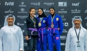 Three female martial artists wearing purple belts and medals stand together on a podium, with two men in traditional attire standing at the sides. The background features logos of event sponsors and organizers.