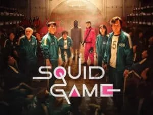 Promotional image for "Squid Game" featuring several characters in green tracksuits standing in a room; a masked figure dressed in black and guards in pink uniforms are in the background. The title "Squid Game" is displayed at the bottom.