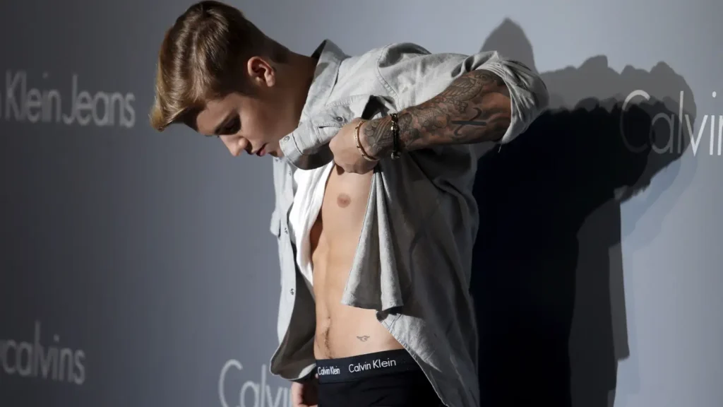 A person lifting their shirt to reveal their torso, standing in front of a Calvin Klein Jeans backdrop, with a tattoo on their arm and wearing Calvin Klein underwear.