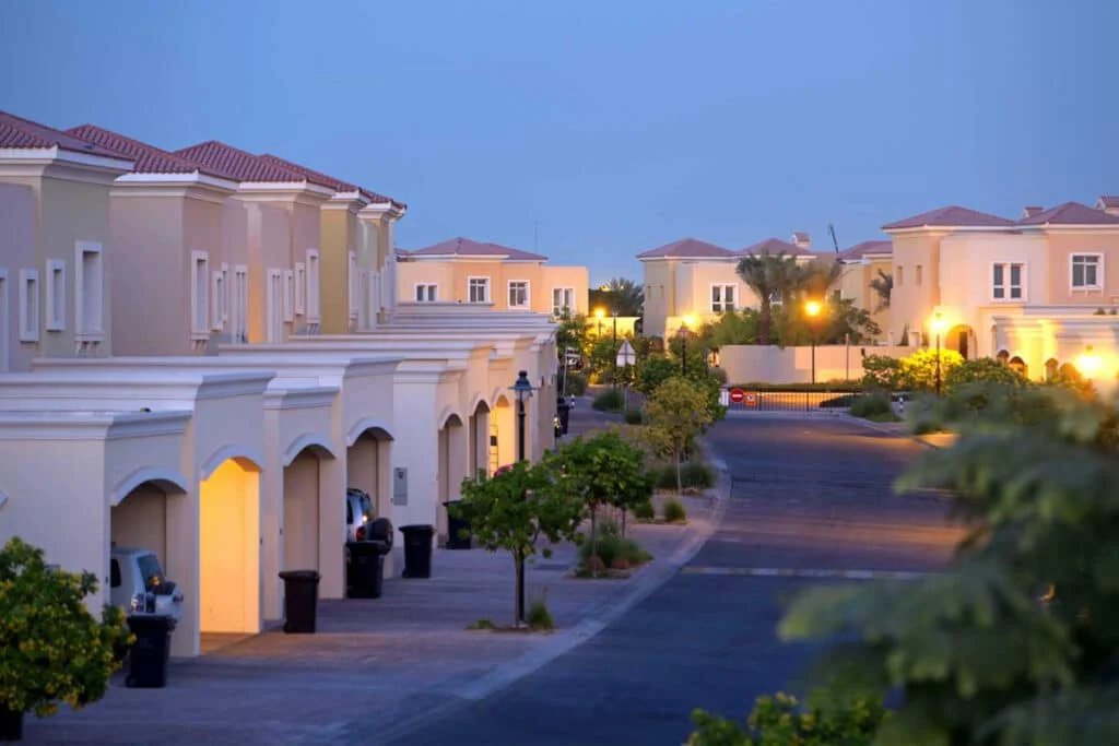 Suburban neighborhood at dusk with rows of beige stucco houses, lit streetlamps, and neatly maintained trees and shrubs along the street.