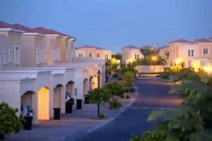 Suburban neighborhood at dusk with rows of beige stucco houses, lit streetlamps, and neatly maintained trees and shrubs along the street.
