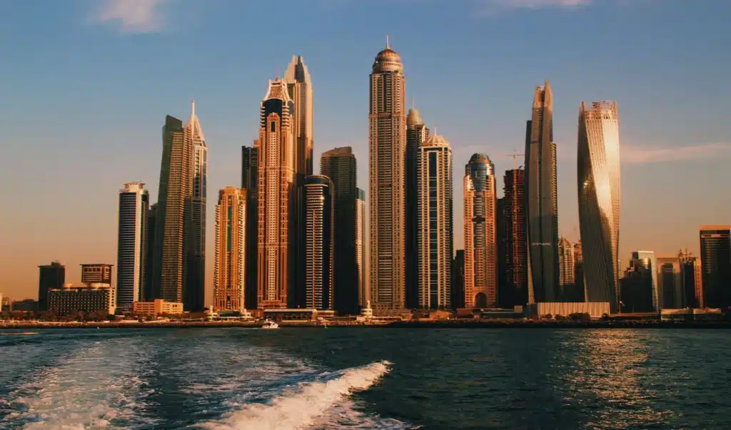 Skyline of a city with tall skyscrapers during sunset, viewed from the water with the wake of a boat visible in the foreground.