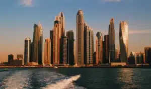 Skyline of a city with tall skyscrapers during sunset, viewed from the water with the wake of a boat visible in the foreground.