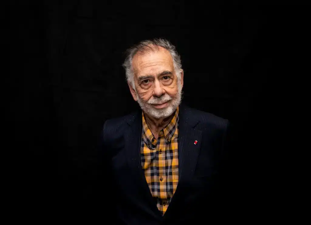 An older man with a beard, wearing a dark pinstripe suit and a yellow plaid shirt, stands against a black background.
