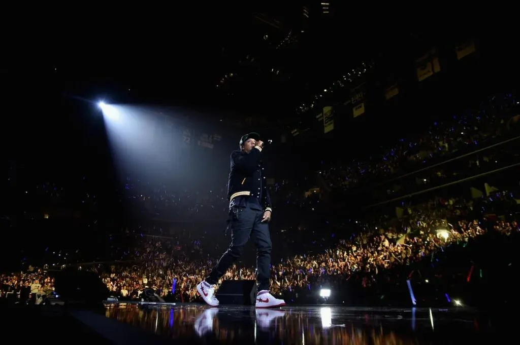 A performer on stage in front of a large audience, illuminated by a spotlight, with vibrant colors from lighting and accessories visible in the crowd.